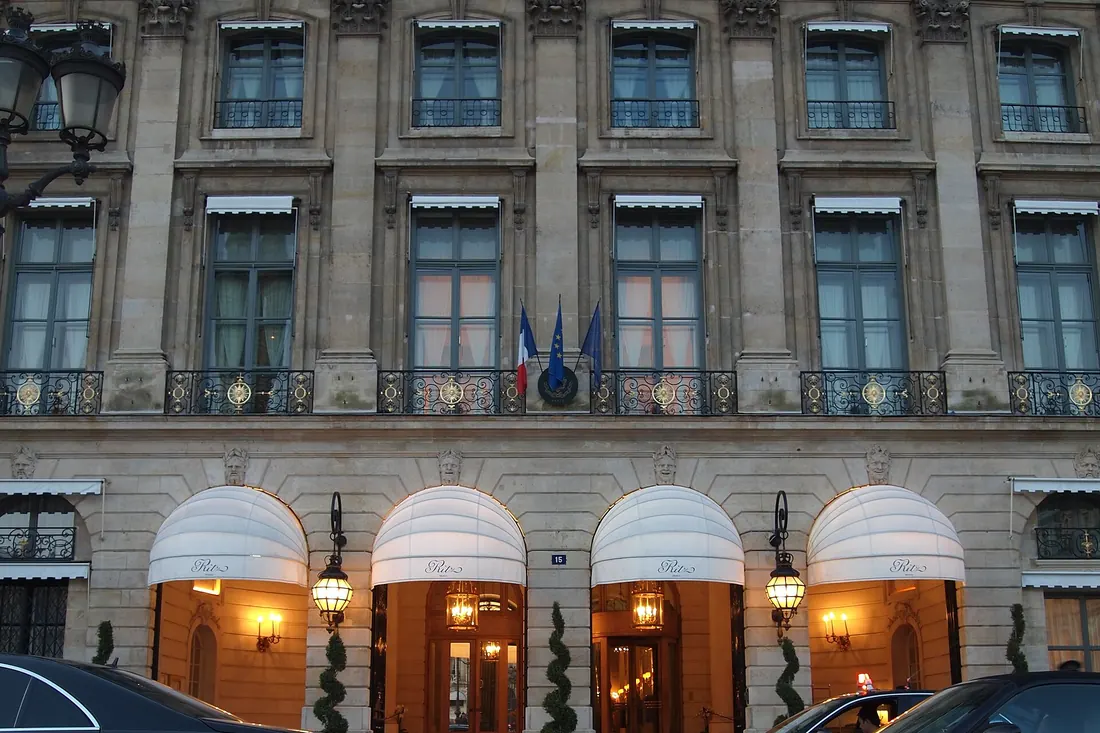Photograph of the Ritz Paris entrance on Place Vendôme: four lit awnings under the stone façade at dusk