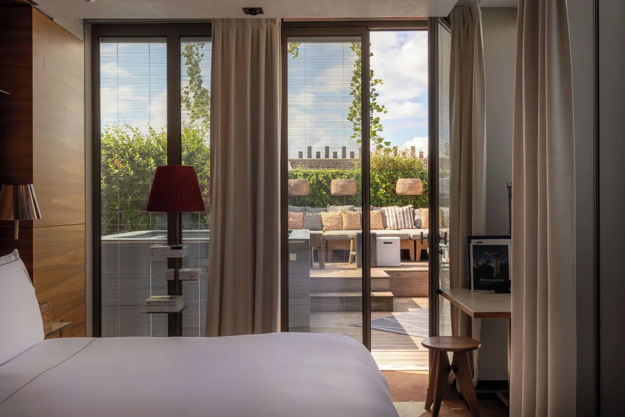 A bed facing French windows open onto a planted terrace with sofas and lanterns