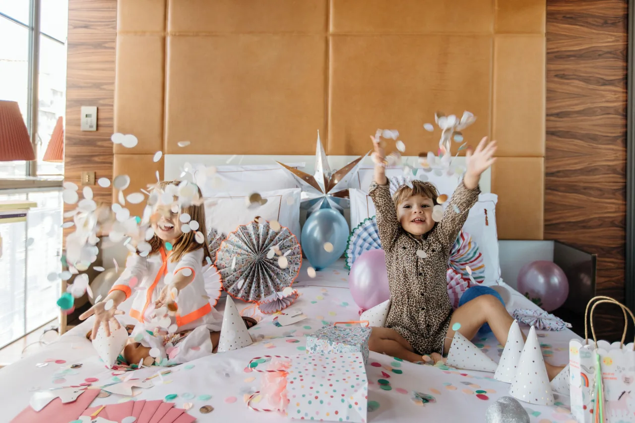 Two children throwing confetti on a bed among balloons and party hats