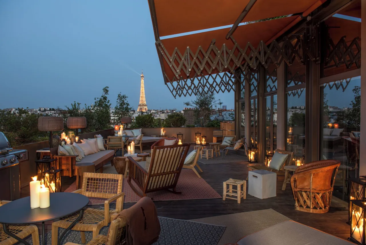 A rooftop terrace at dusk with candles and armchairs under a striped awning, the lit Eiffel Tower in the distance