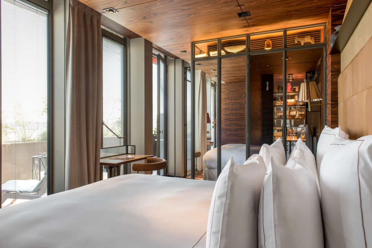 A bedroom with a white bed, tall windows with curtains and wooden shelves