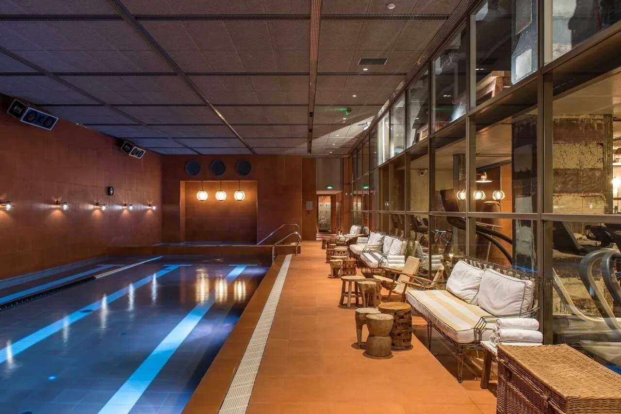 A long indoor pool with lit lanes, loungers along a glass wall and warm wood-panelled walls