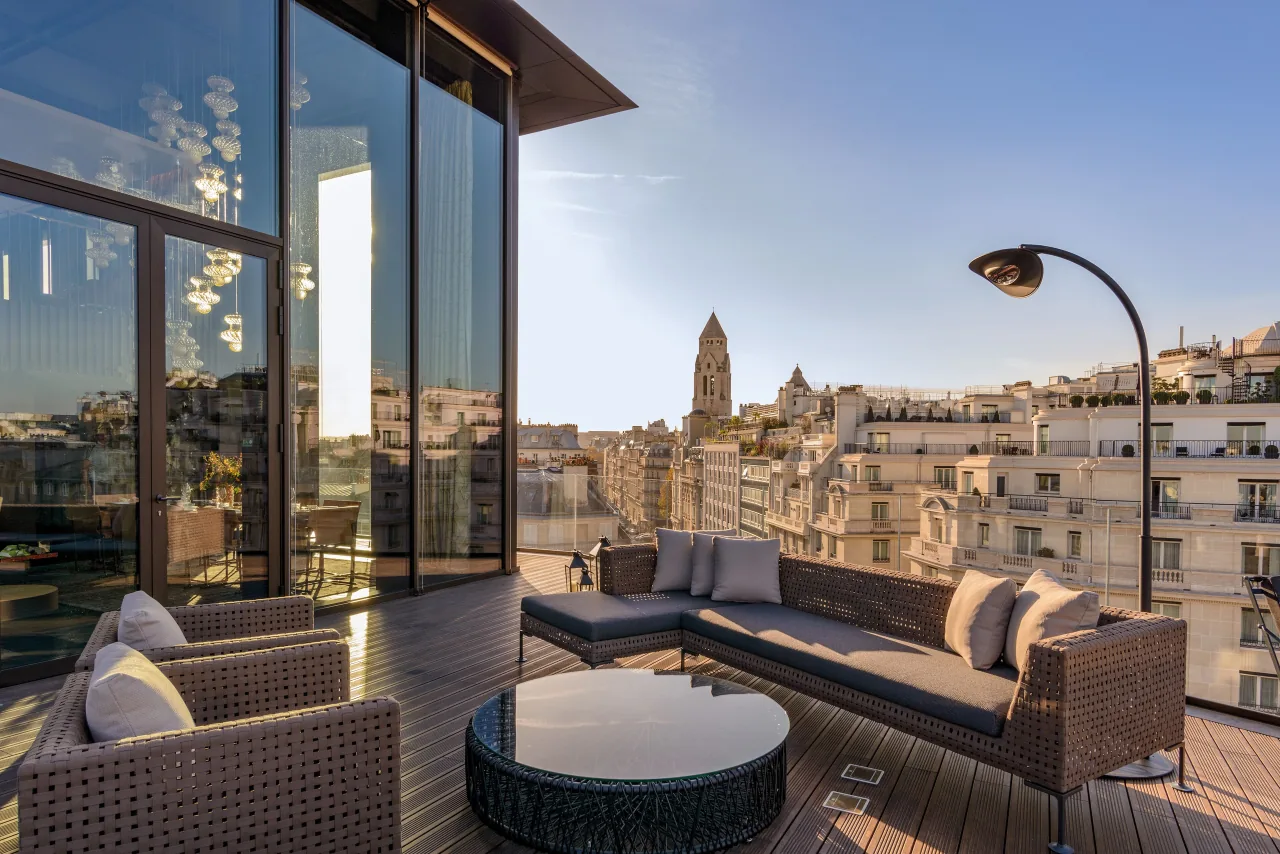 A high terrace with wicker sofas, a round table and a lamp, over Paris rooftops and a church bell tower