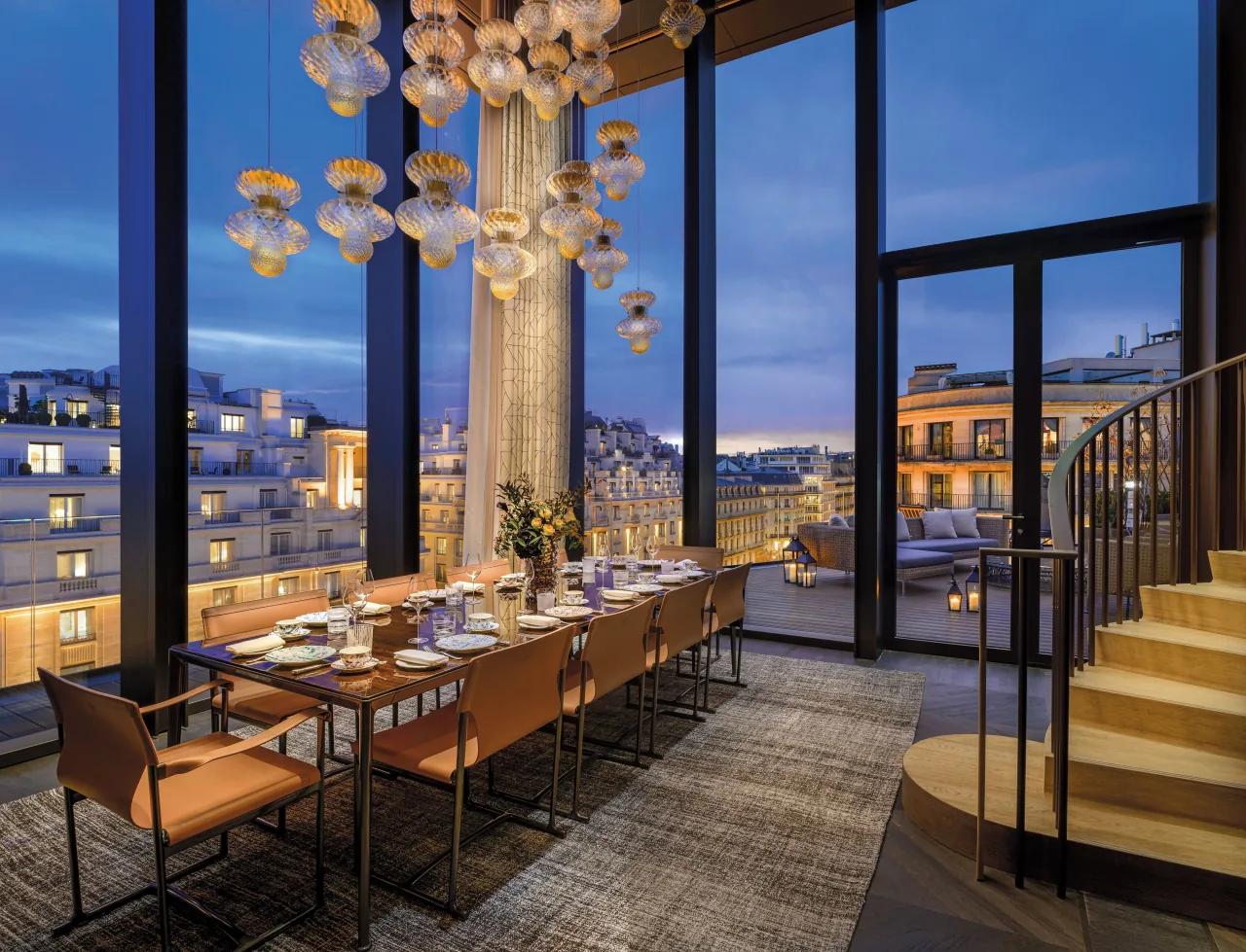 A glass-walled dining room at dusk, a long table under a glass chandelier and a staircase, over the rooftops