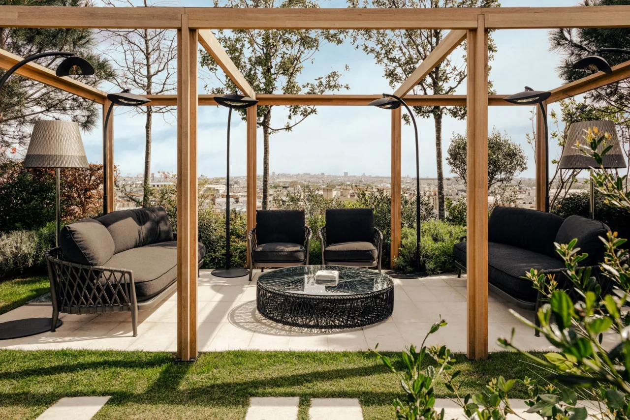 A wooden pergola over outdoor sofas and a round table, on a lawn among trees