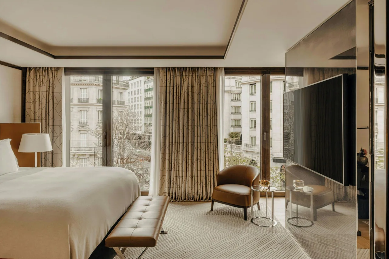 A bedroom with a bench at the foot of the bed, curtained floor-to-ceiling windows and a television on a stand