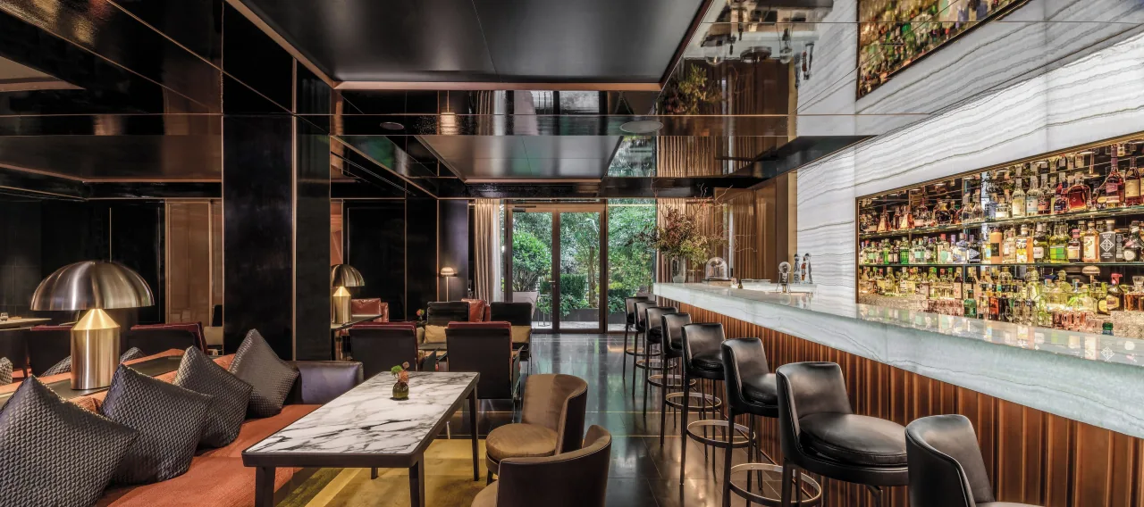 A bar with a long pale counter, a wall of bottles and leather armchairs
