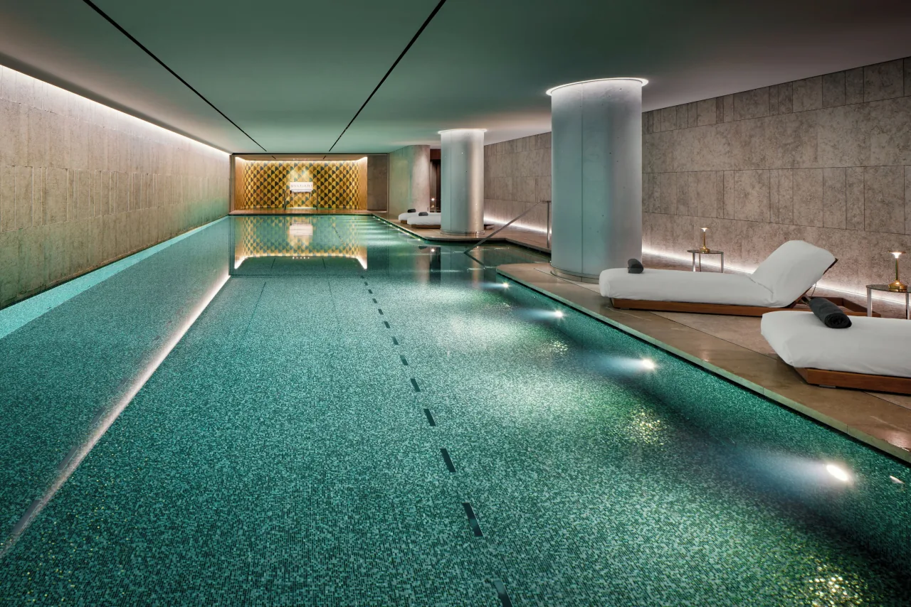 A long indoor pool with green mosaics, white loungers along one side and glowing columns