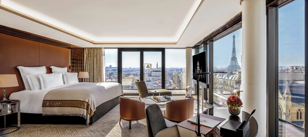 A bedroom with a large bed, armchairs and a round column, the Eiffel Tower visible through the windows