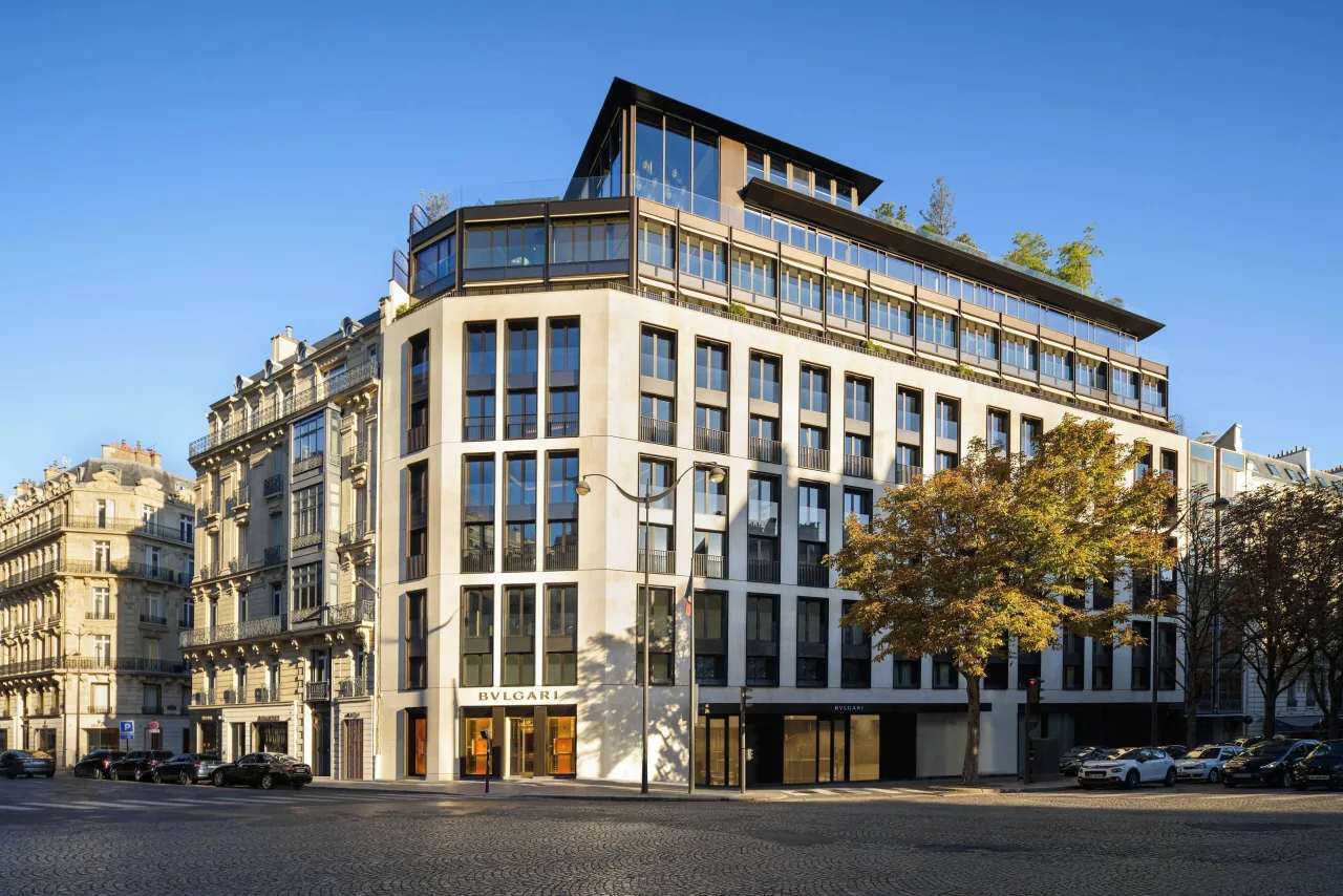 A pale stone building on a street corner, its upper floors glazed and planted, under a blue sky