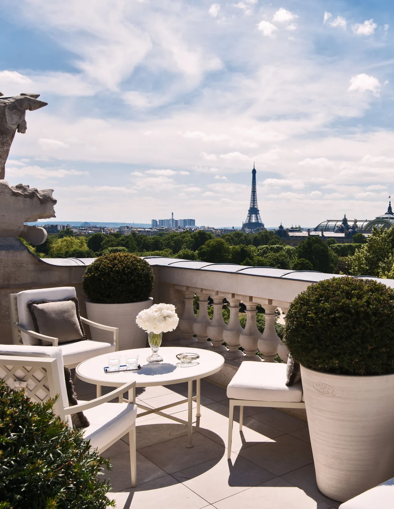 Une terrasse à balustrade de pierre, chaises blanches et buis taillés, au-dessus des arbres, la tour Eiffel au loin