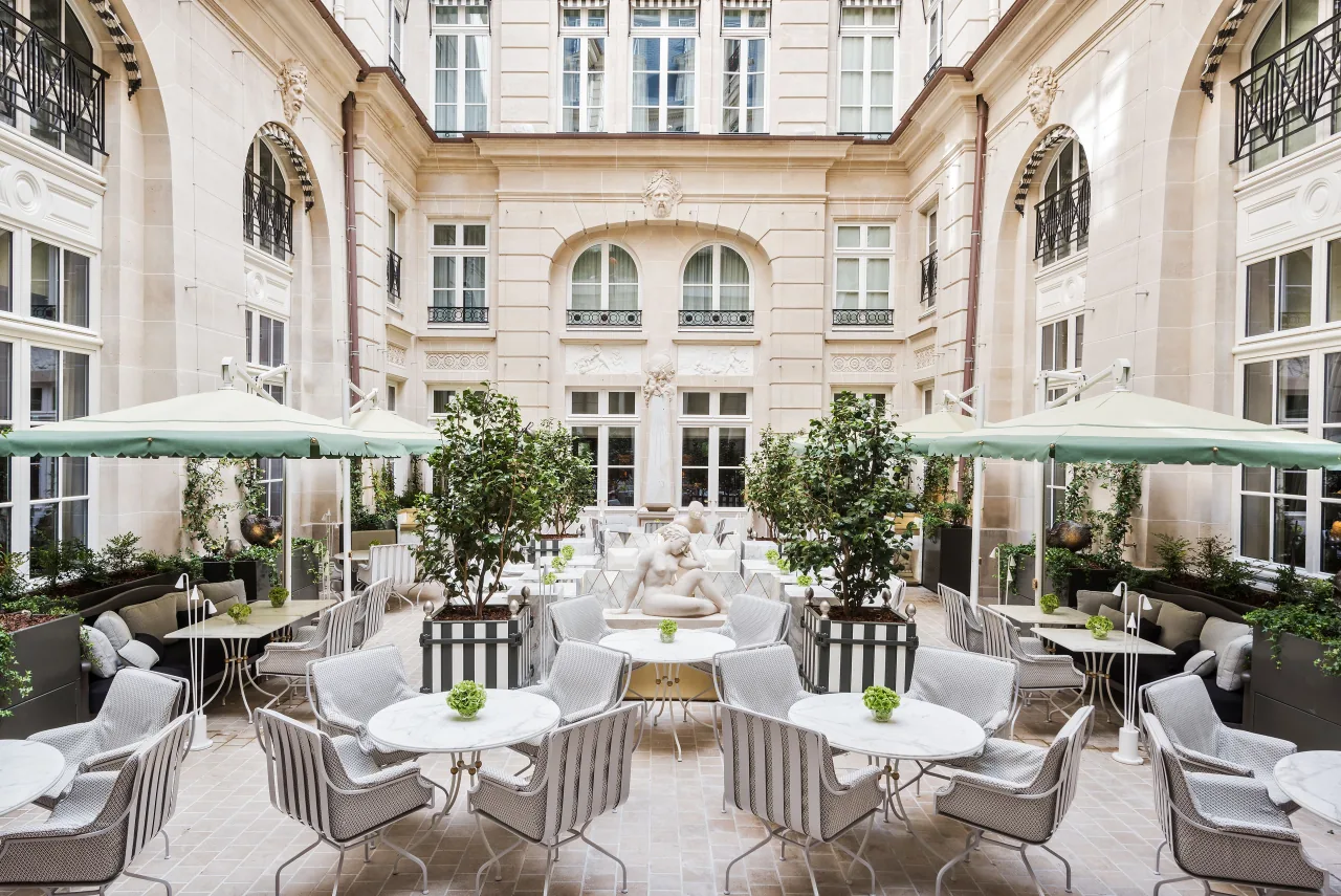 Une cour intérieure de pierre claire aux fenêtres cintrées, garnie de tables et de fauteuils blancs, de parasols verts et d’oliviers en bac