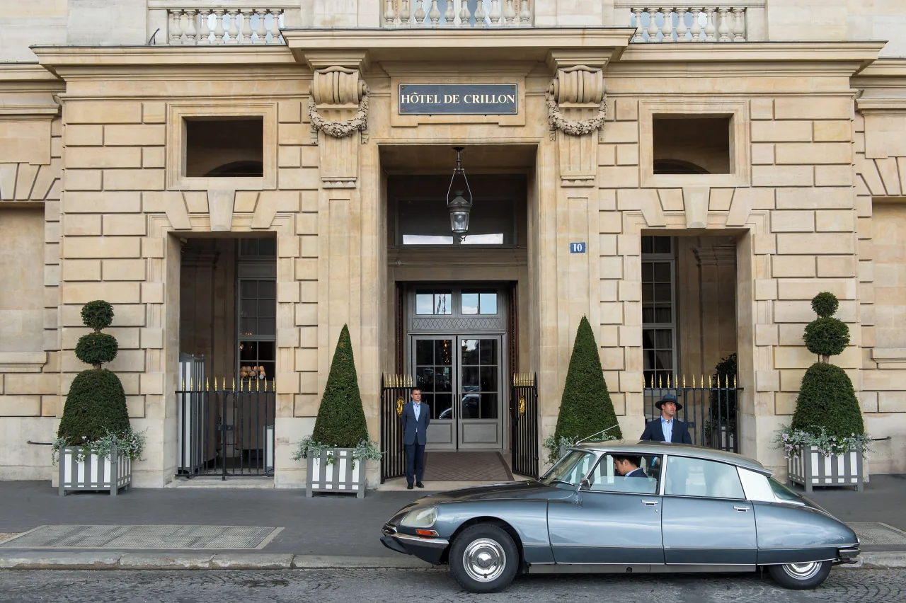 Une entrée de pierre avec une lanterne, des topiaires taillés en bac et une voiture ancienne argentée garée devant la porte