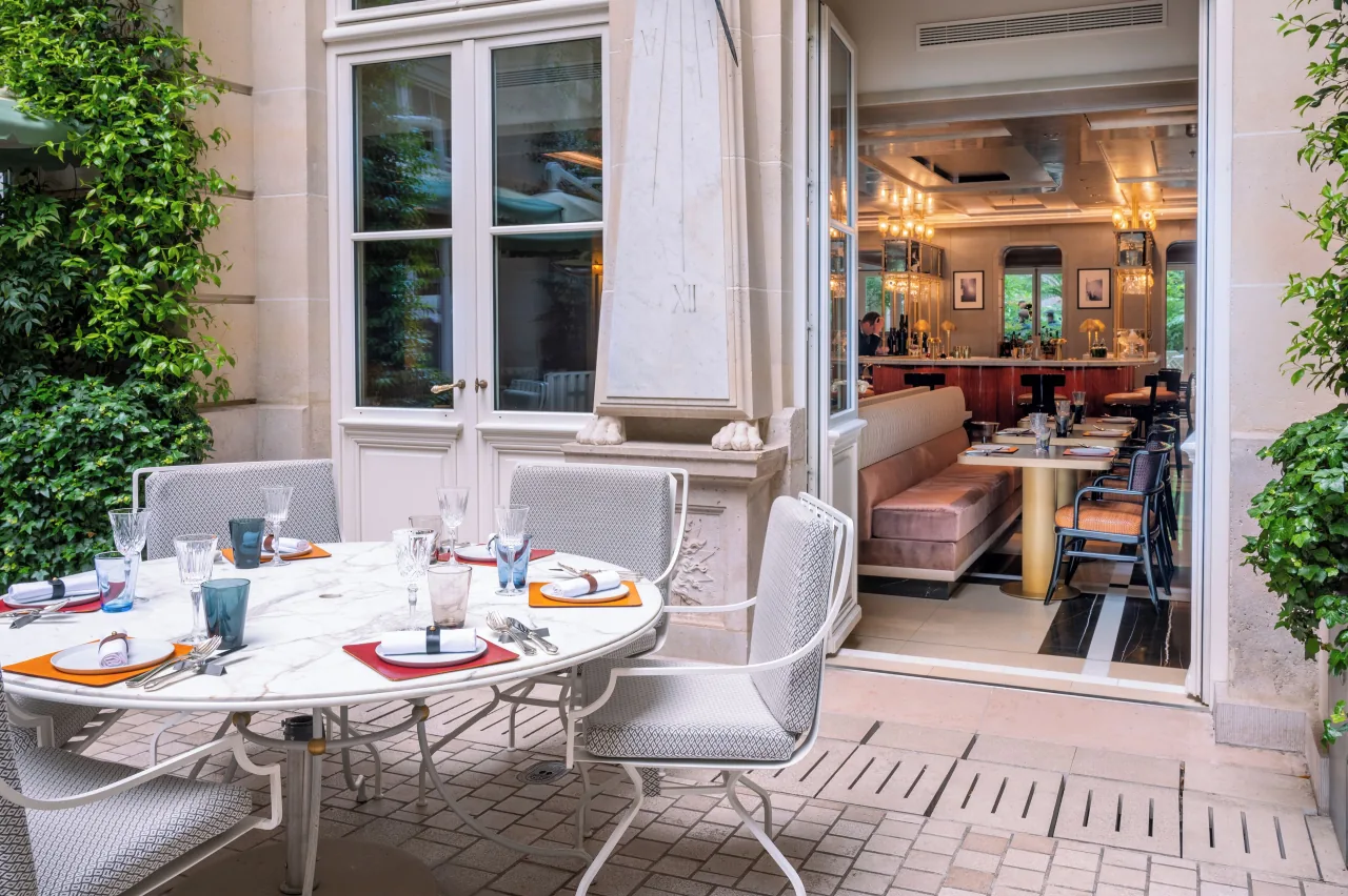 Une table dressée sur une terrasse de cour, devant les portes ouvertes d’un restaurant à banquettes