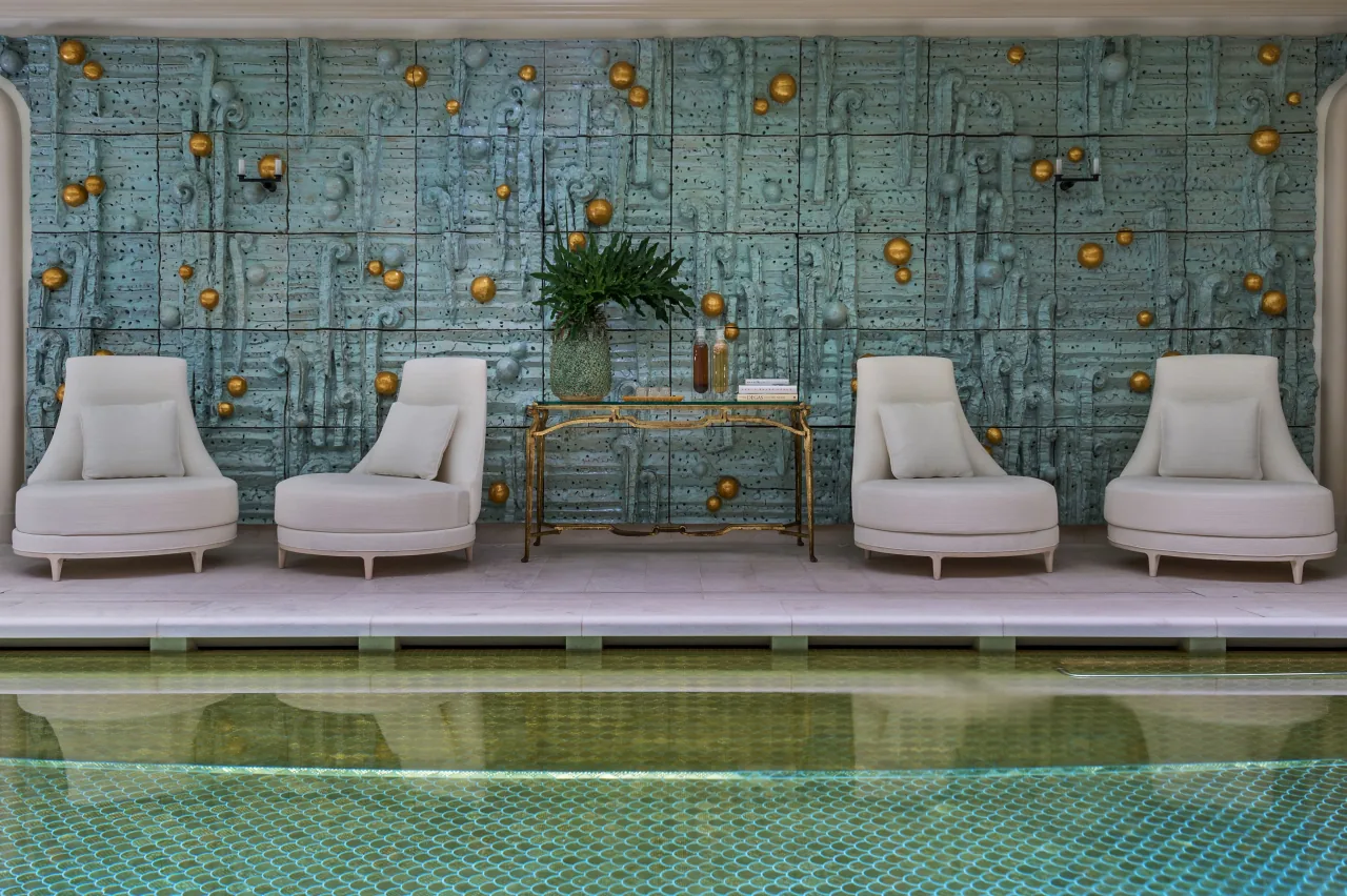 Une piscine intérieure au fond de mosaïque, des transats blancs au bord et un mur orné d’une fresque de céramique bleue