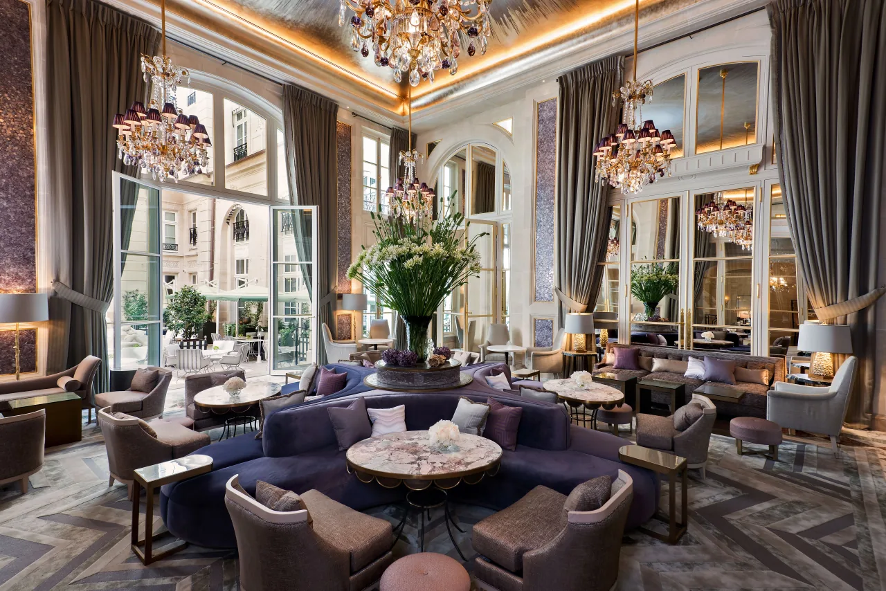 Un haut salon aux lustres de cristal, fauteuils et canapés de velours, tables rondes et grandes fenêtres