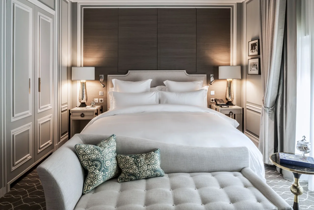 Une chambre, un lit blanc, une banquette capitonnée, des coussins à motifs et une tête de lit sombre entre deux lampes
