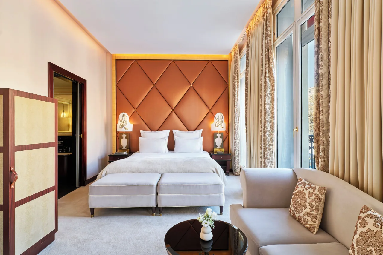 Une chambre, une tête de lit capitonnée cuivrée, un canapé clair et de hautes fenêtres à rideaux