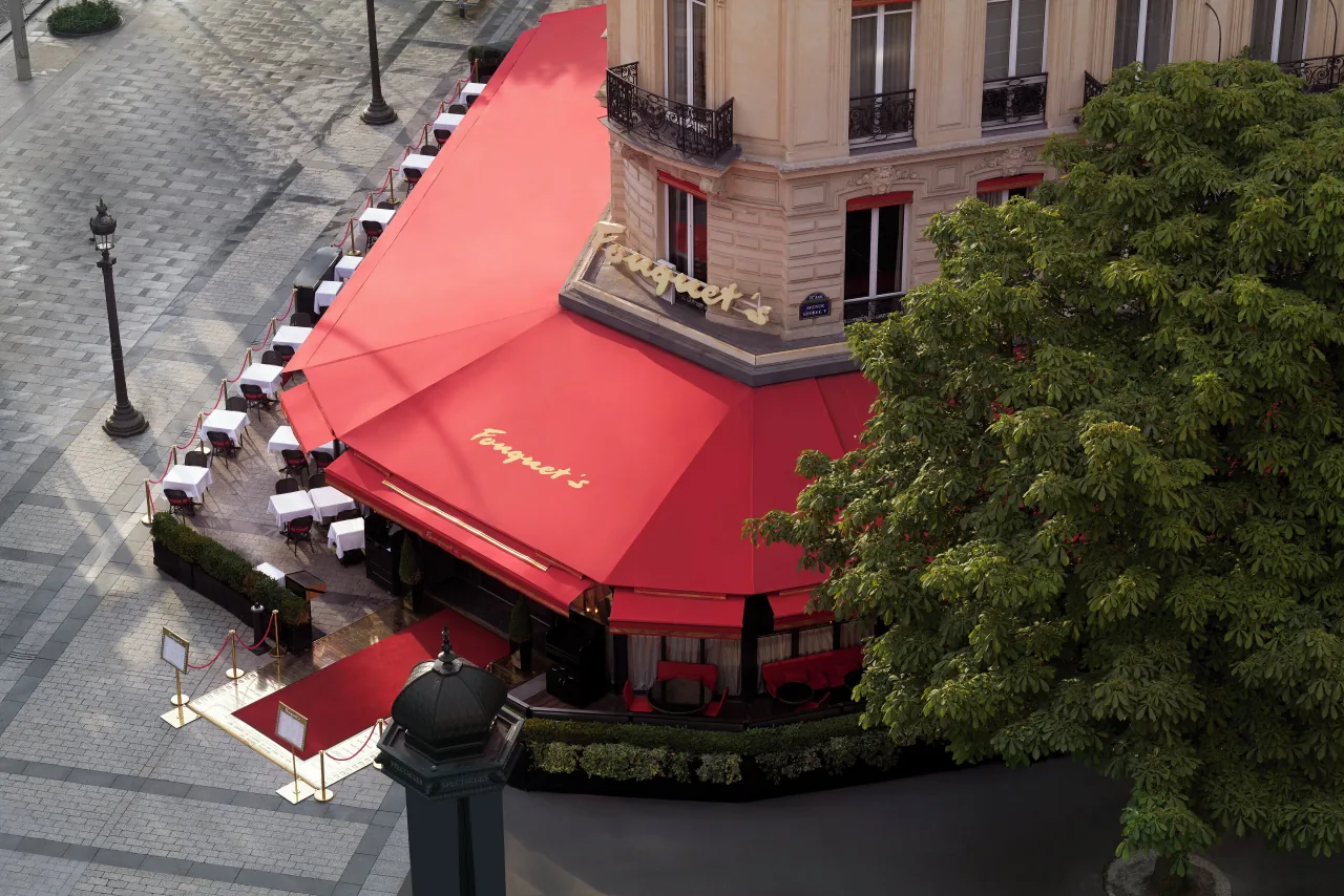 Un angle de rue vu d’en haut, des auvents rouges à lettres d’or au-dessus d’une terrasse, un réverbère et des arbres
