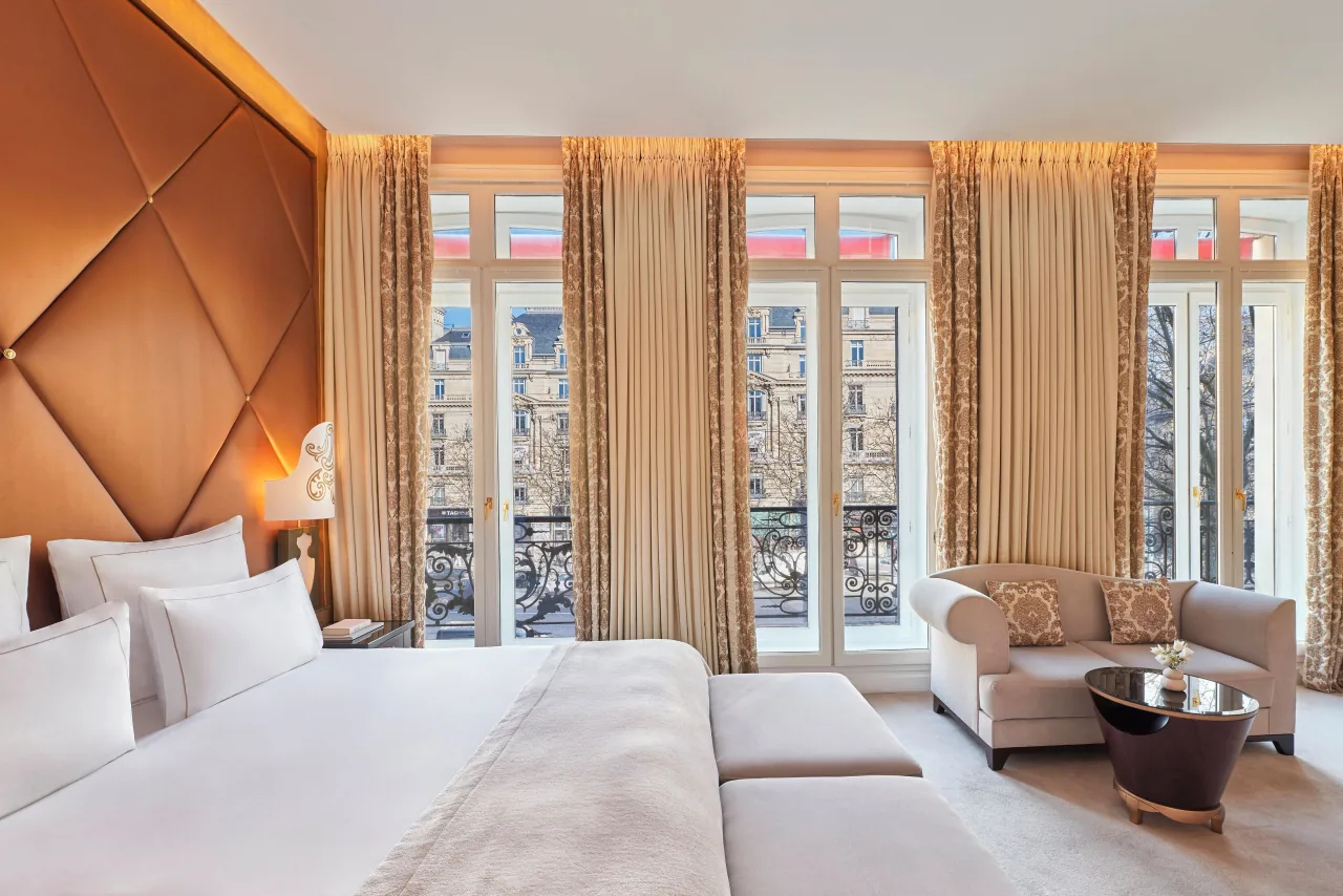 Une chambre lumineuse, un grand lit à tête de lit capitonnée, un fauteuil et de hautes fenêtres