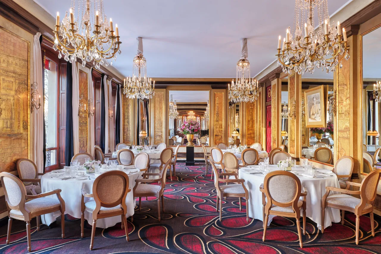 Une salle à manger, des tables rondes nappées de blanc, des lustres de cristal, des murs dorés et un tapis rouge à motifs
