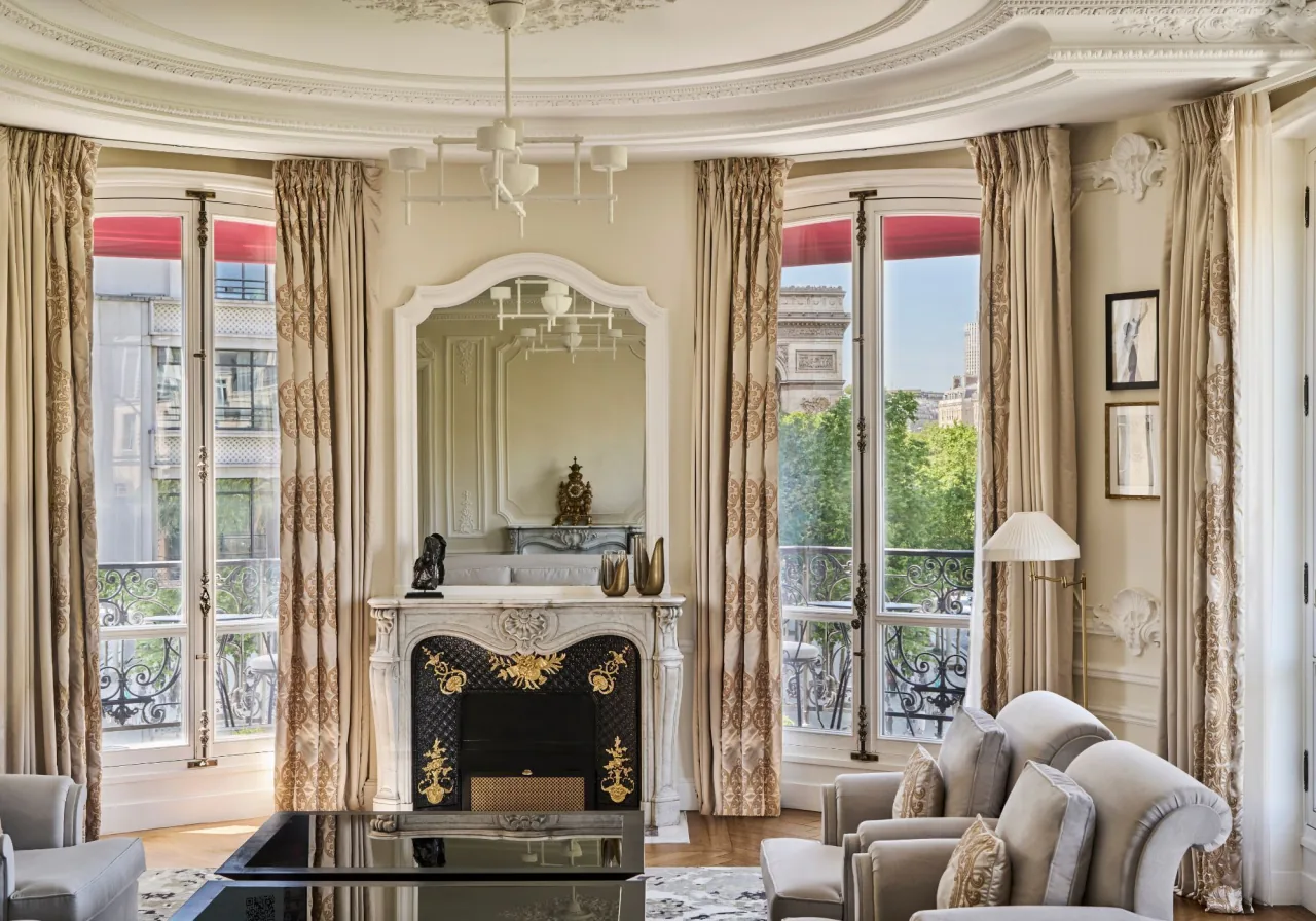Un salon, une cheminée de marbre, un miroir doré, des canapés clairs et de hautes fenêtres sur un balcon et des arbres