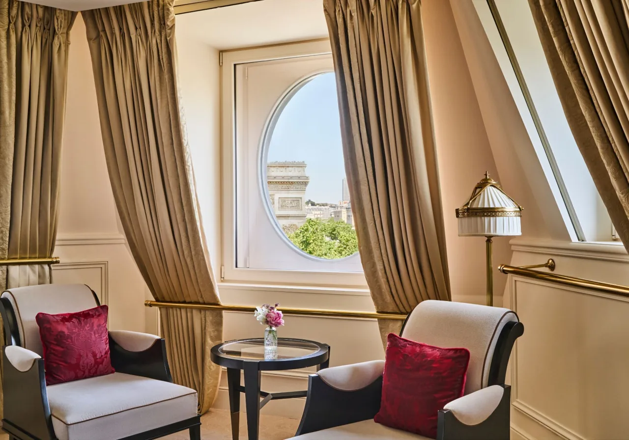 Une fenêtre ronde sous les combles qui cadre l’Arc de Triomphe, deux fauteuils à coussins rouges et une petite table