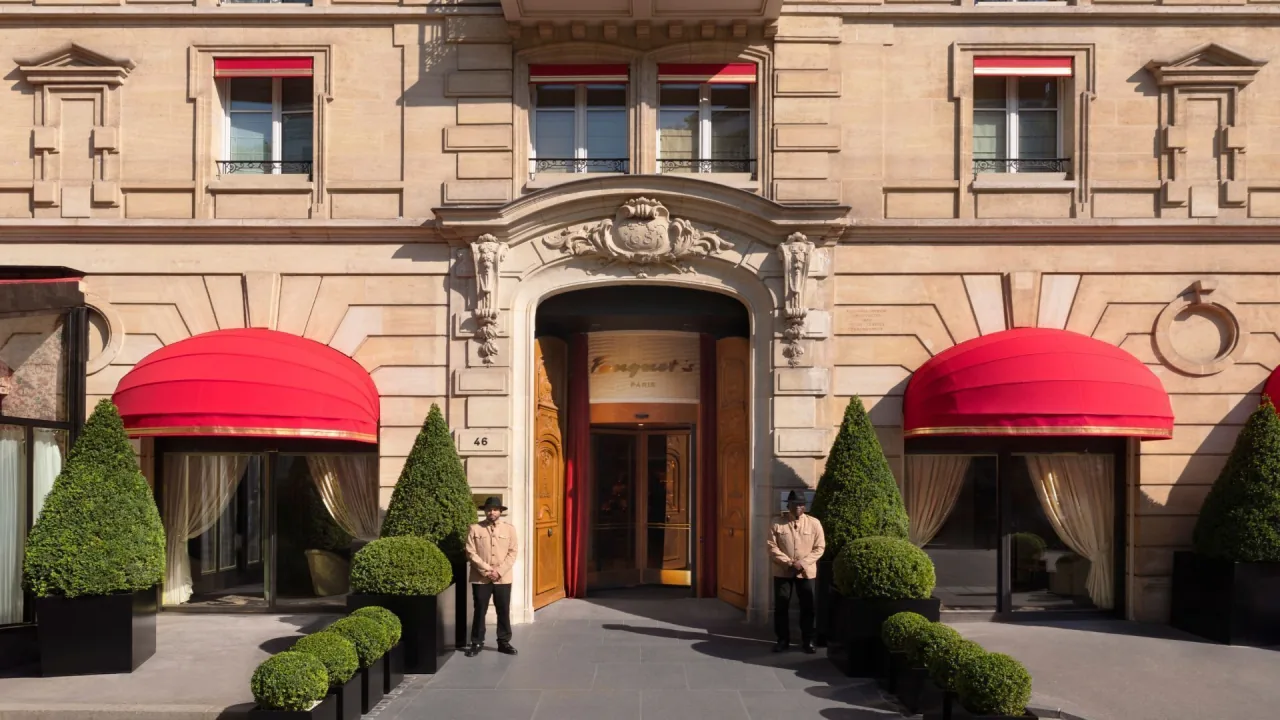 Une façade de pierre, une entrée cintrée entre deux auvents rouges, des arbustes taillés et deux portiers