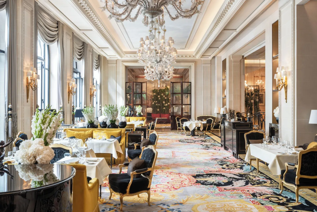 Une grande salle à manger, lustres de cristal, hautes fenêtres, fauteuils jaunes et tapis à motifs