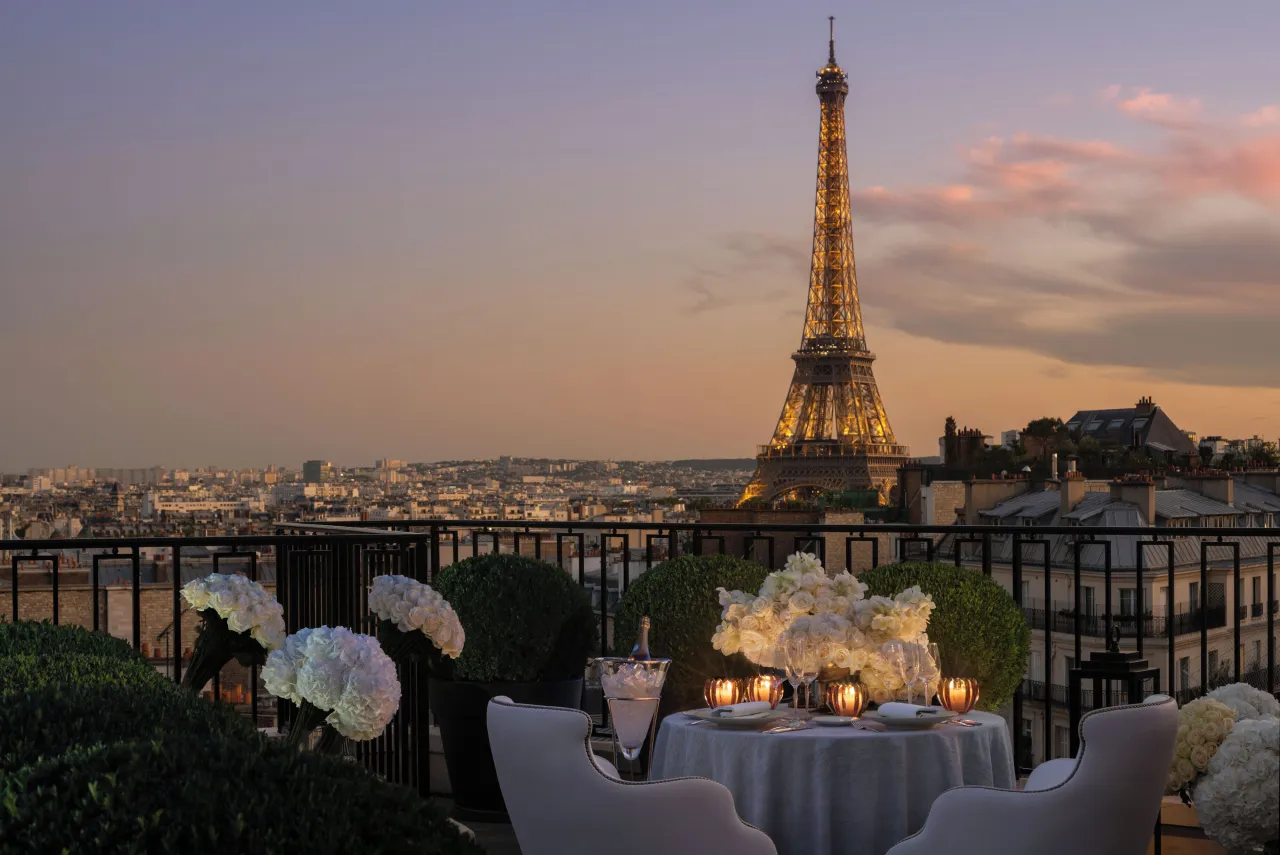 Une table dressée de bougies et de fleurs blanches sur une terrasse au crépuscule, la tour Eiffel illuminée au-dessus des toits