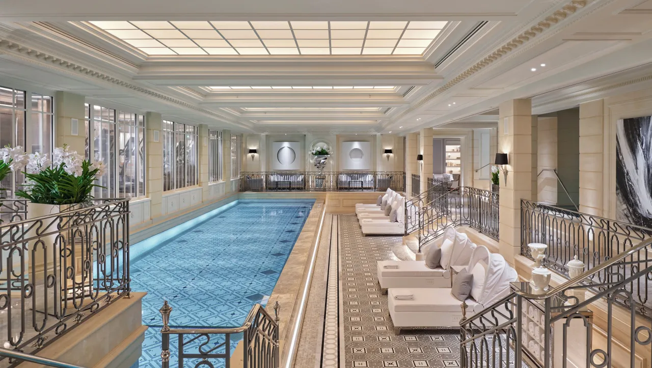 Une longue piscine intérieure en mosaïque bleue sous un plafond vitré, rambardes en fer forgé et chaises longues blanches