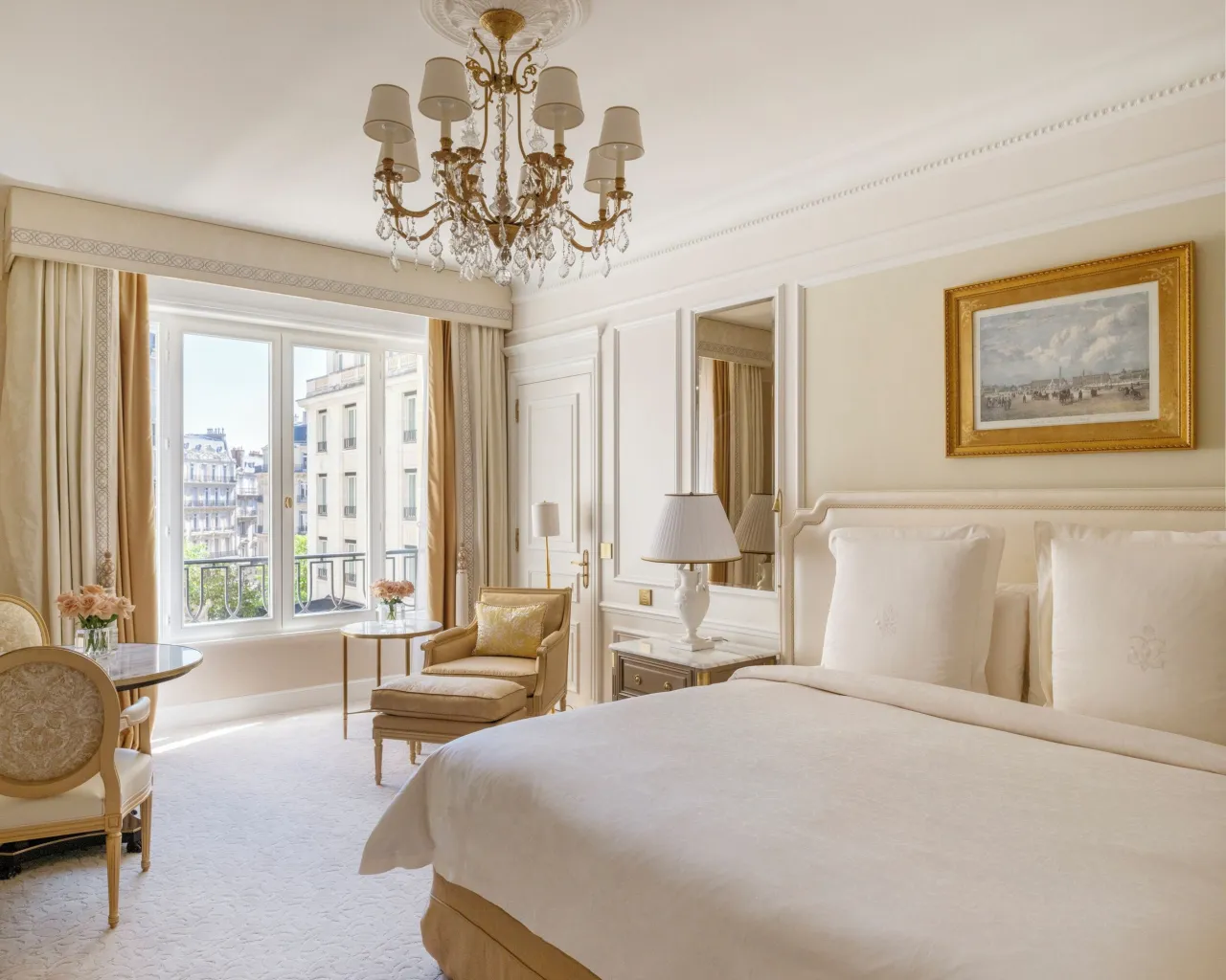 Une chambre aux tons crème, lustre, grand lit blanc, tableau et porte-fenêtre sur l’avenue