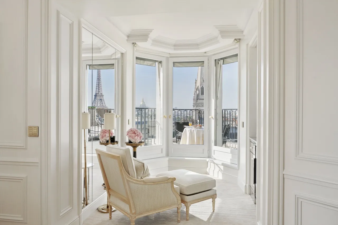 Un coin salon blanc, fauteuil et repose-pieds dans une fenêtre en rotonde, la tour Eiffel et une flèche d’église au loin