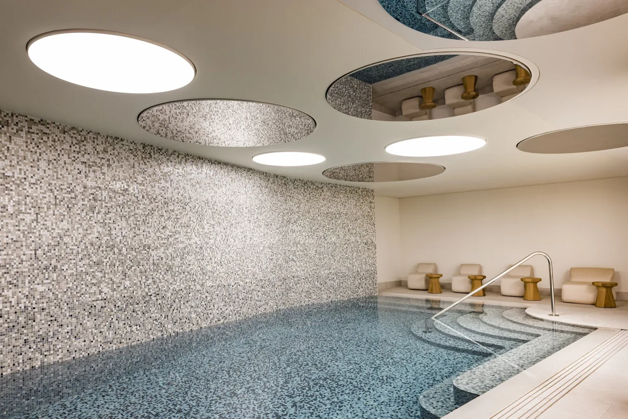 Une piscine intérieure en mosaïque gris-vert sous des luminaires ronds, avec des marches et des transats