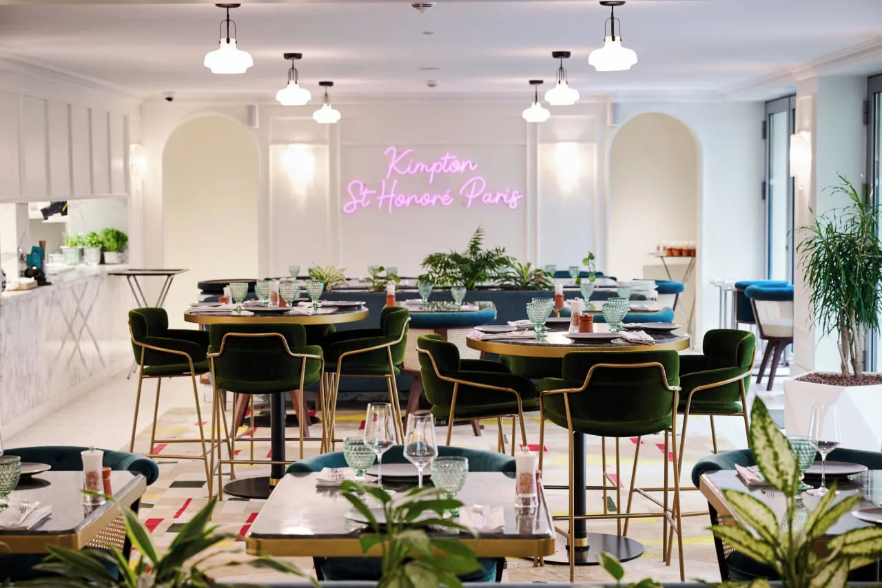 Une salle à manger claire, des chaises vertes, des tables rondes, des plantes et une enseigne au néon rose