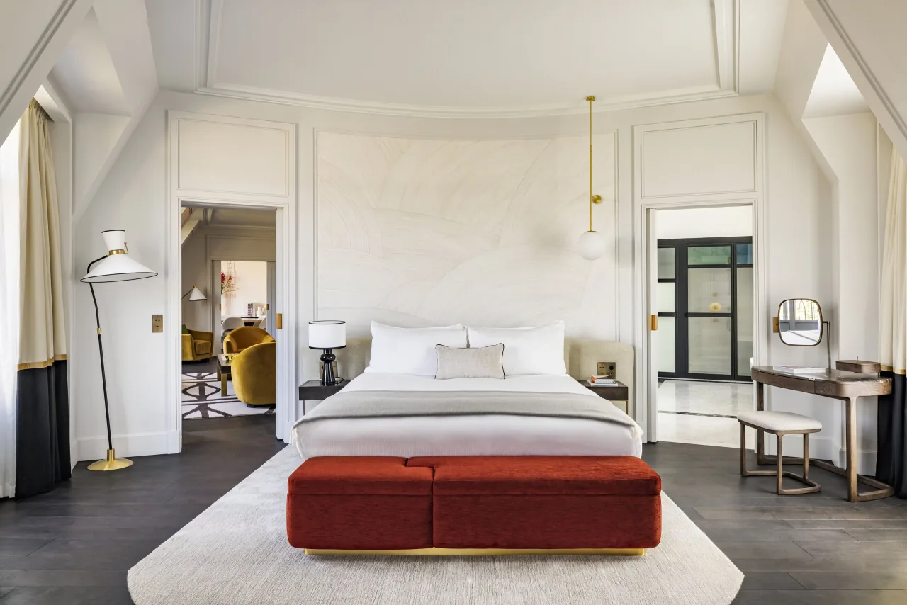 Une chambre blanche, un banc de velours rouge au pied du lit et des portes ouvertes sur la pièce voisine