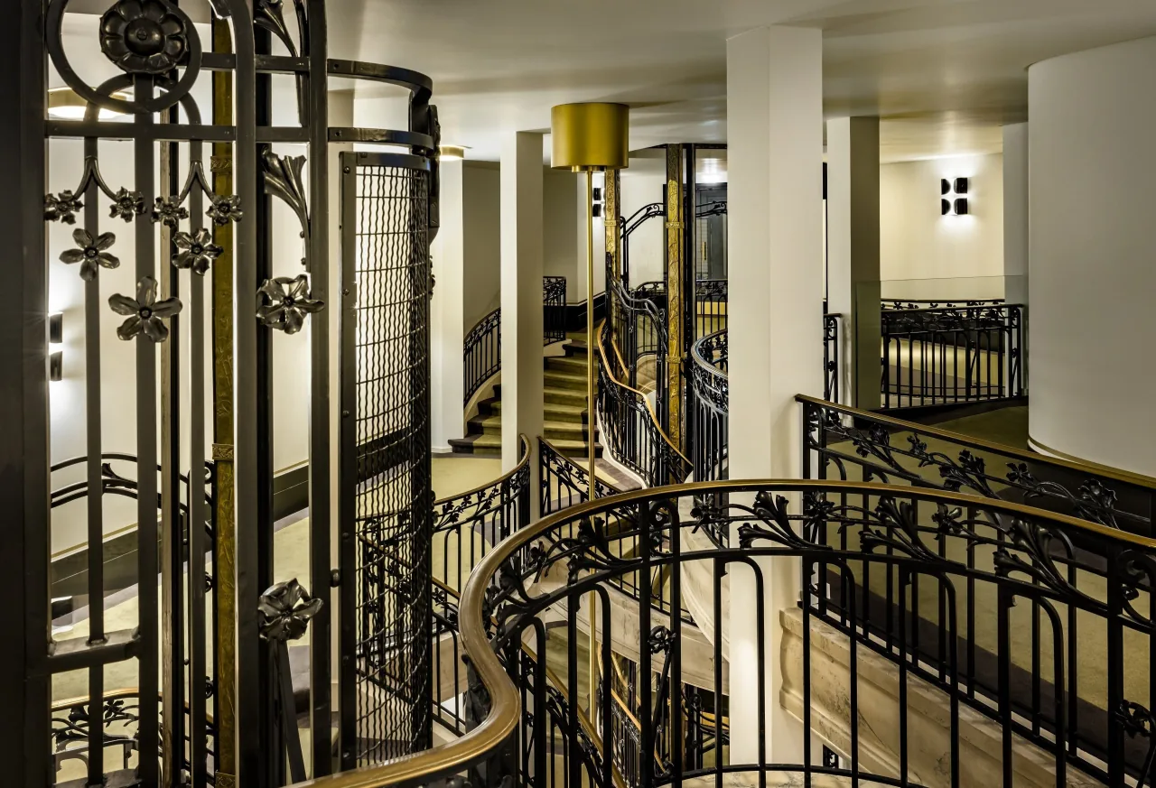 Une cage d’ascenseur en fer forgé à côté d’un escalier courbe aux rampes dorées
