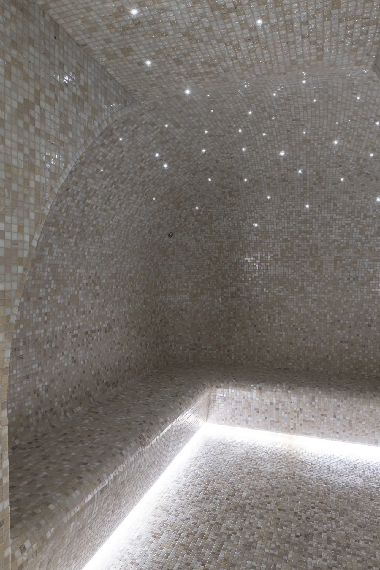 Un hammam en mosaïque gris clair, un banc éclairé et de petites lumières au plafond