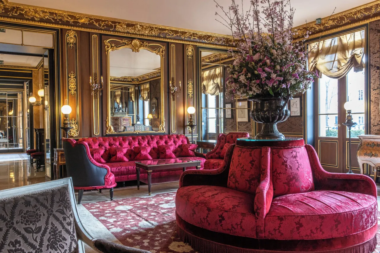Un salon aux boiseries dorées et aux miroirs, des canapés de velours rouge et une banquette ronde sous un grand vase de fleurs