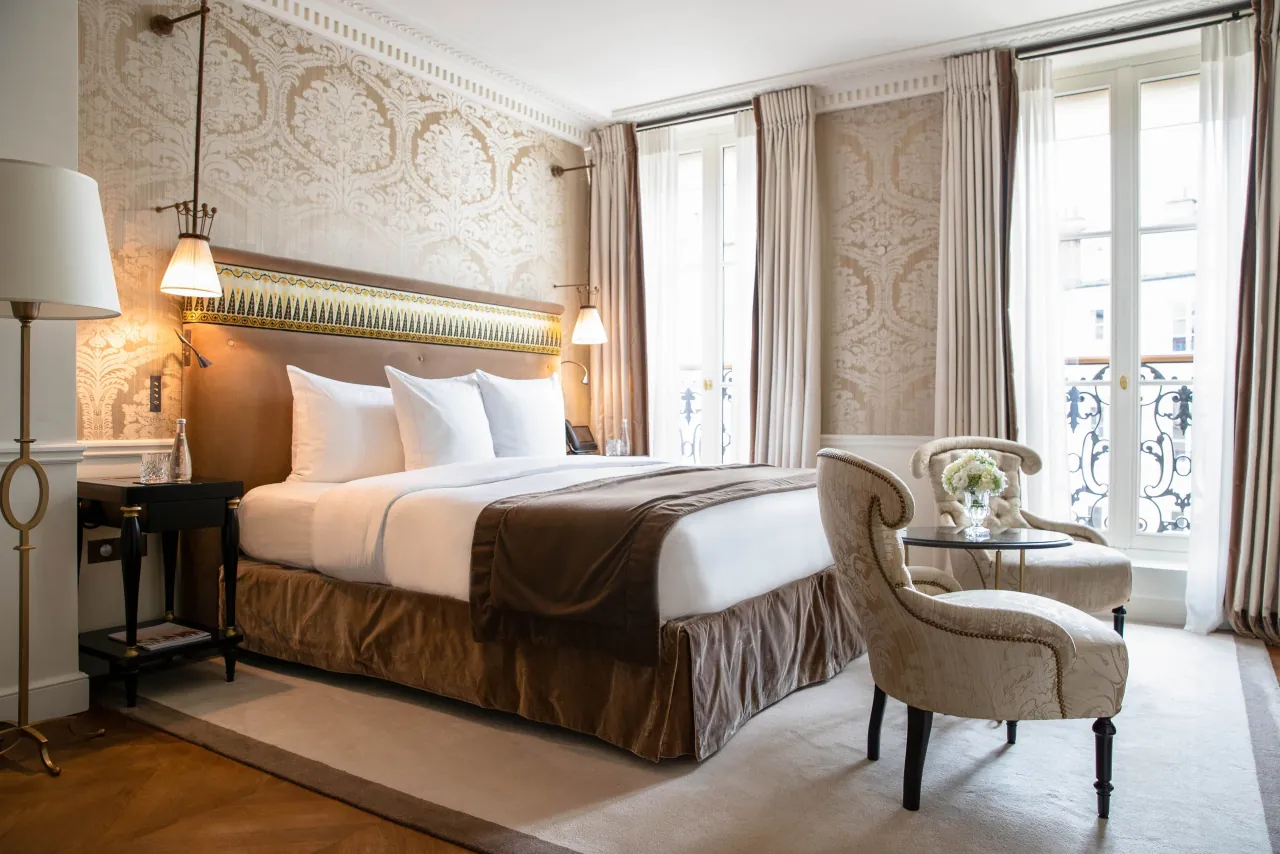Une chambre, une tête de lit capitonnée, un fauteuil clair et des portes-fenêtres sur un balcon en fer forgé