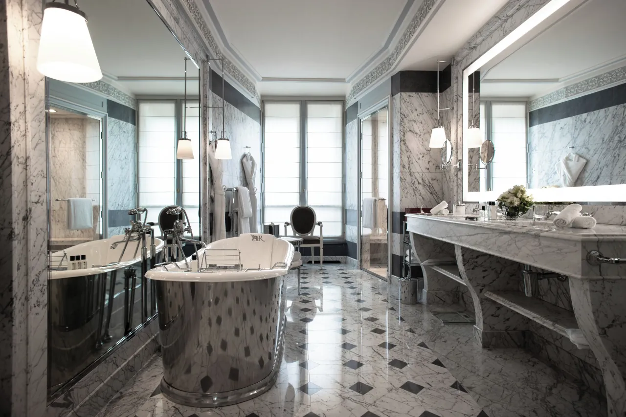 Une salle de bains en marbre gris, une baignoire sur pieds en métal, une double vasque et une chaise rouge