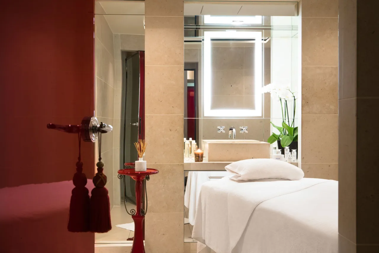 Une cabine de soins, une table de massage, une porte rouge à poignée à pompons et un miroir éclairé