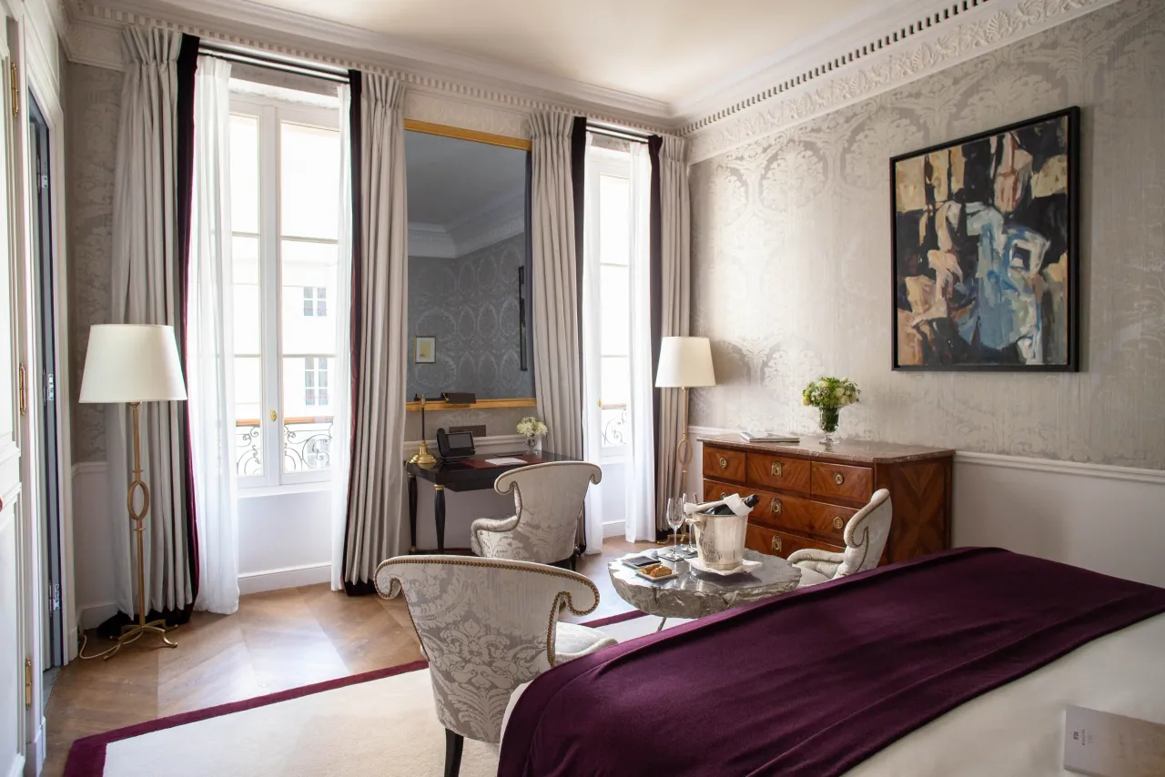 Une chambre, un plaid prune, une commode dorée, un tableau abstrait et de hautes fenêtres