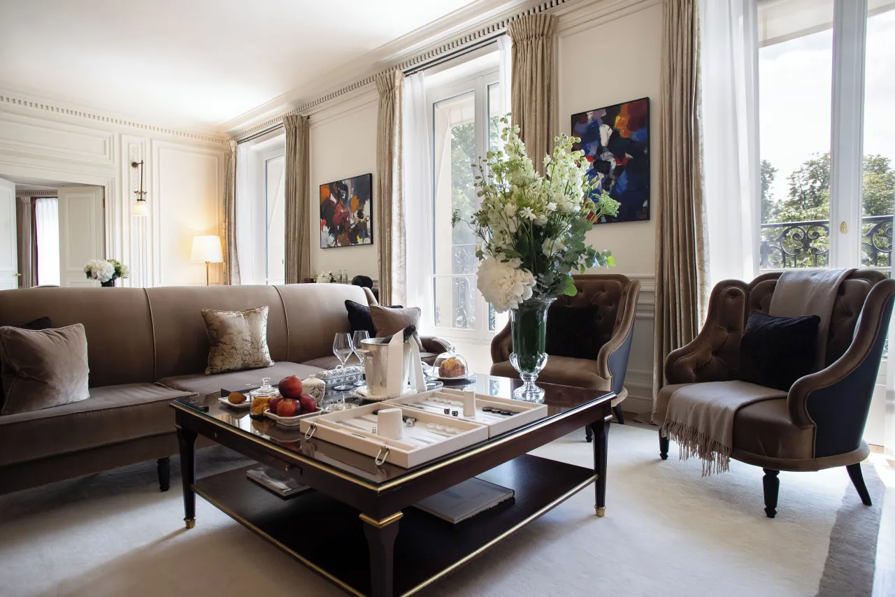 Un salon, des canapés et fauteuils de velours, une table basse garnie de fruits, des fleurs et des tableaux contemporains