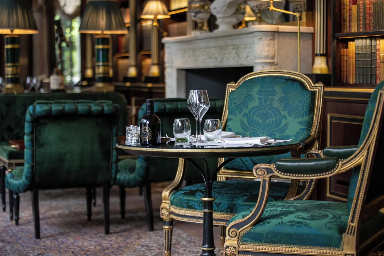 Une table dressée de verres entre des fauteuils de velours vert, une cheminée et des rayonnages derrière