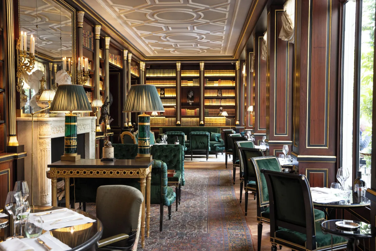 Une bibliothèque aux boiseries, des rayonnages, une cheminée de marbre et des fauteuils de velours vert autour de guéridons