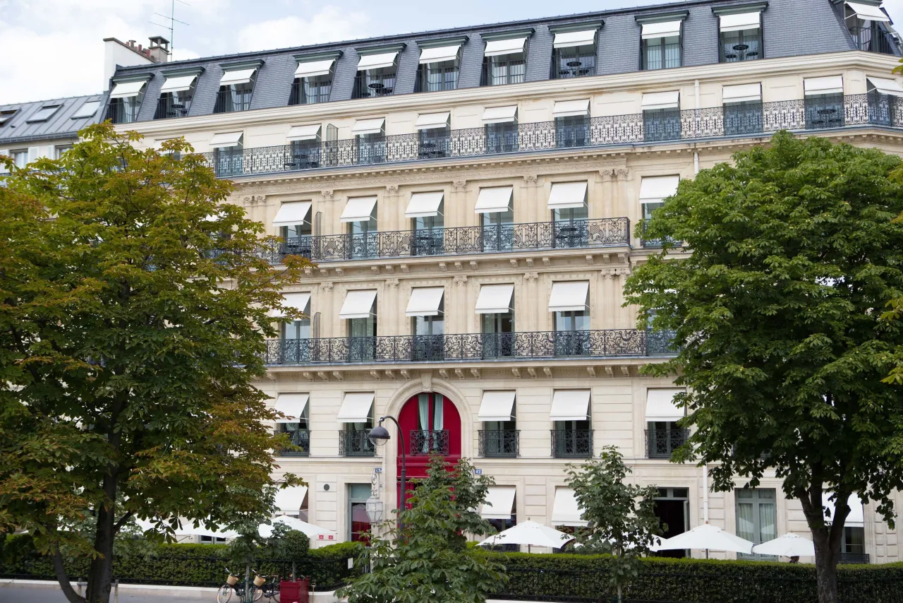 Une façade de pierre claire, des balcons en fer forgé, des stores rayés et une porte rouge cintrée entre deux arbres