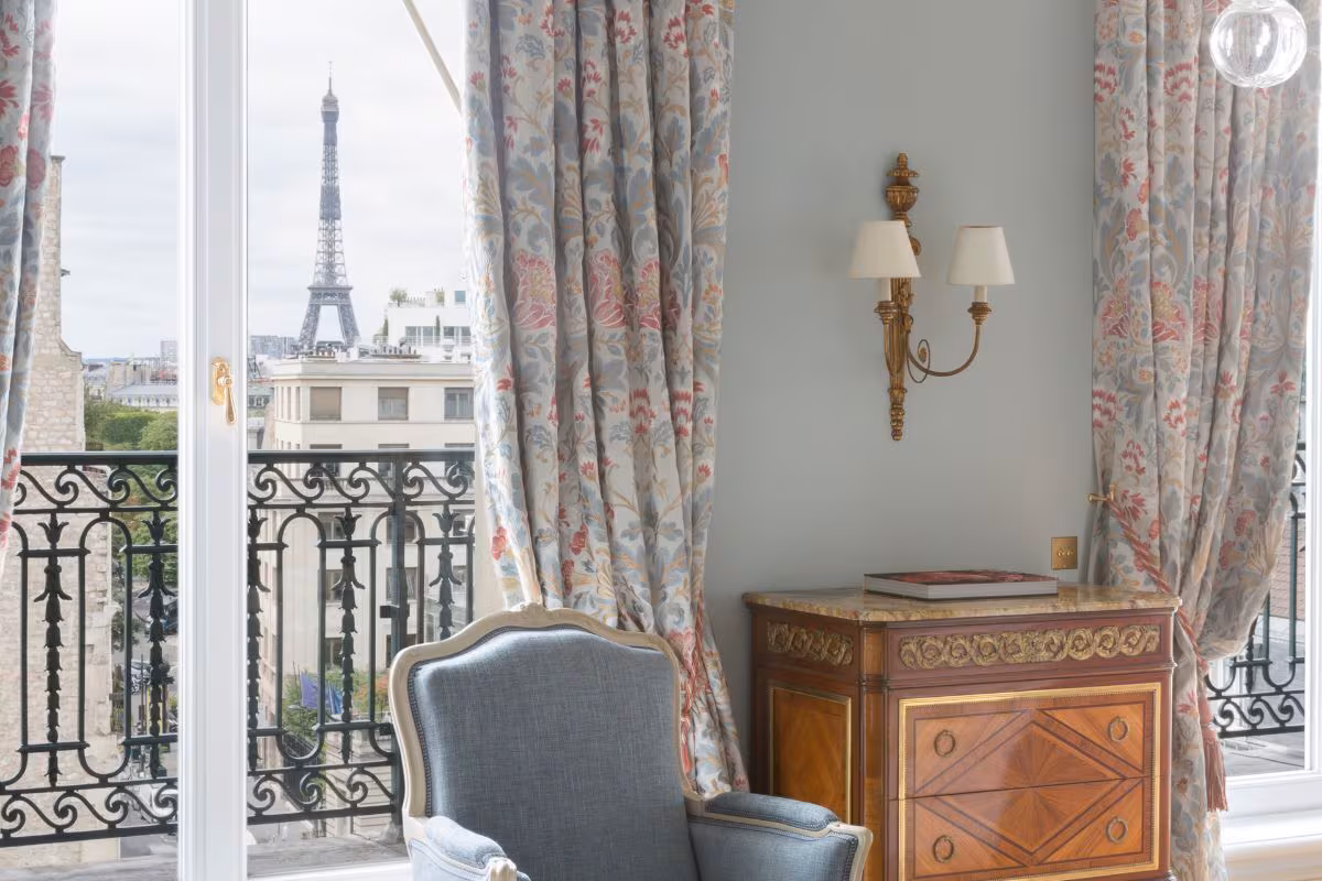Vue sur la tour Eiffel depuis une chambre du Bristol Paris, par une porte-fenêtre à balcon en fer forgé, entre rideaux fleuris