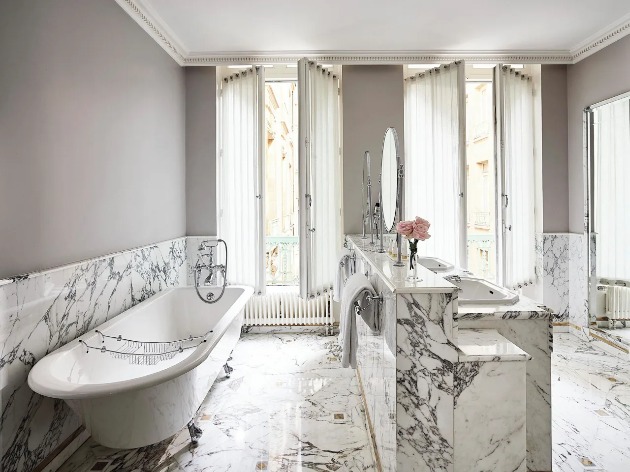 Une salle de bains de marbre blanc, une baignoire sur pieds, une double vasque et de hautes fenêtres voilées
