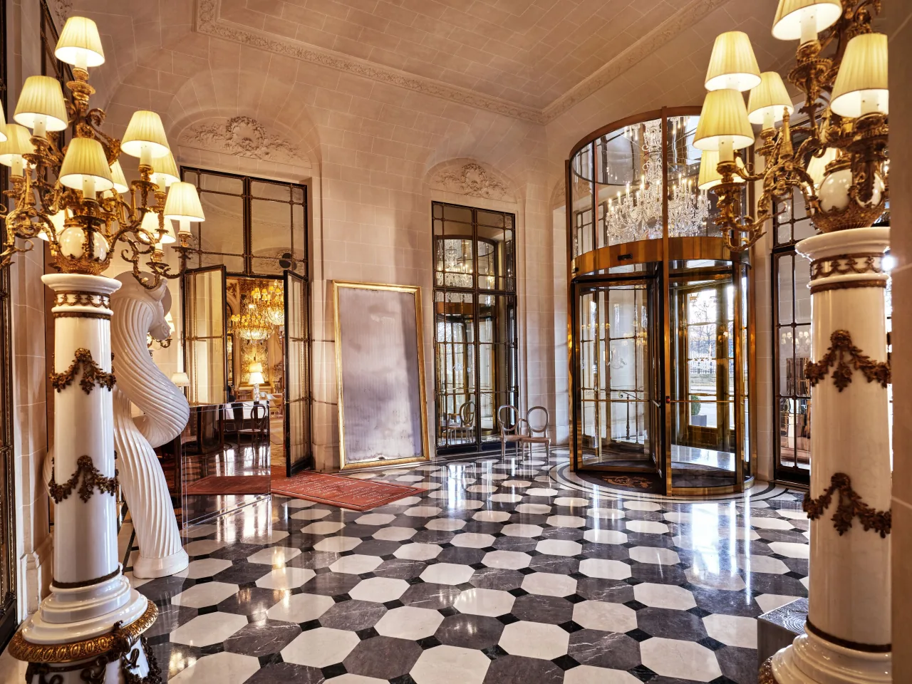 Un hall au sol de marbre noir et blanc, des colonnes blanches sculptées de guirlandes, des lustres dorés à abat-jour et une porte à tambour en laiton